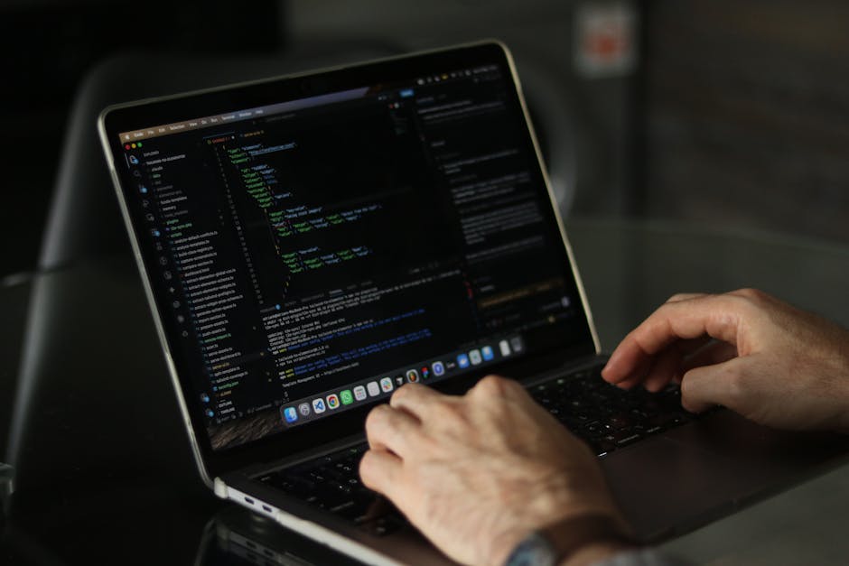 A person working on a laptop with a black screen displaying lines of colorful code and text, situated on a dark table indoors. The individual's hands are visible, with one hand positioned on the keybo