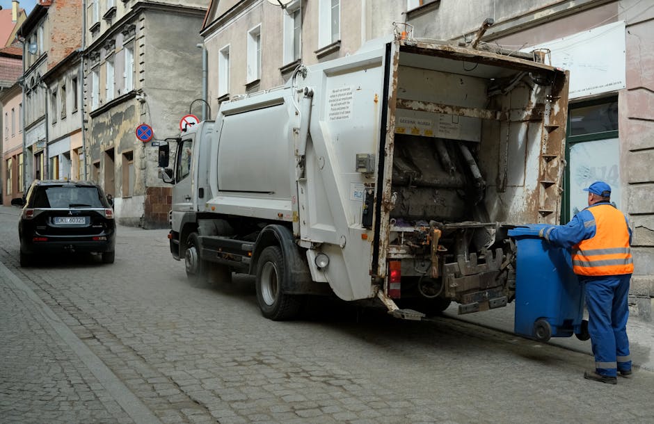 Two large black plastic garbage bags and a cardboard box filled with waste are placed on the edge of a paved street near a metal fence, which has vertical bars and a dark finish. The bags appear crump